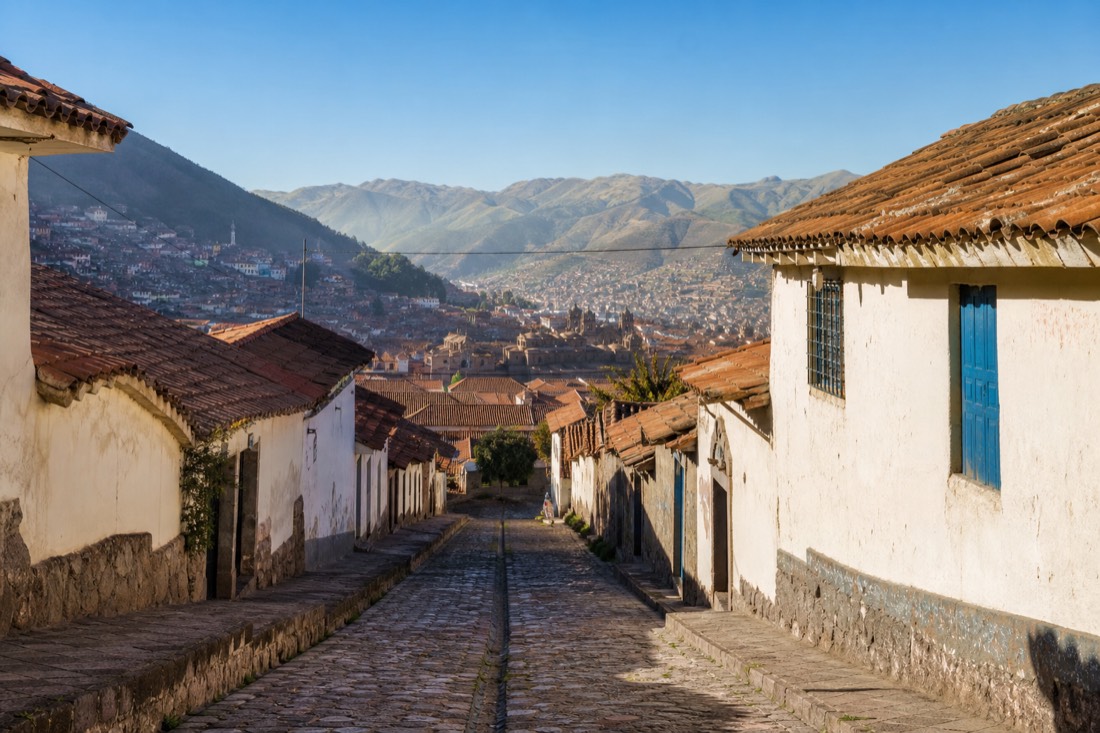 Cobbled Cusco street climbing toward terracotta rooftops with distant green Andean hills under a clear morning sky, no people
