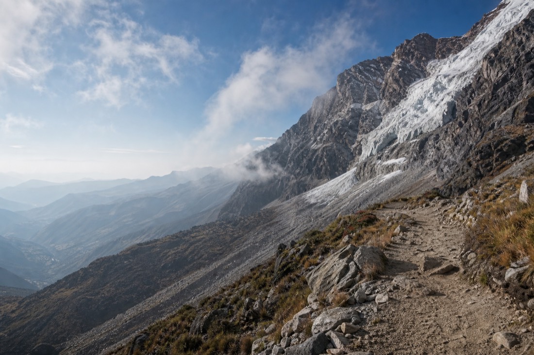 High rocky trail winding up toward a hanging glacier with the valley falling away below in early morning light, empty of people