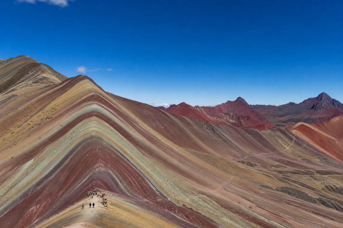 Striped mineral bands of Rainbow Mountain under a deep blue sky with hikers on the ridge trail