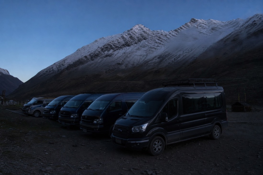 Row of tour vans parked at the Soraypampa trailhead before dawn, snow peaks faint against a dark blue sky