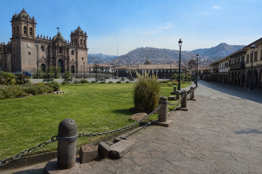 Cusco's historic plaza busy with dry-season travellers under clear Andean light, colonial arches and distant hills, no readable faces