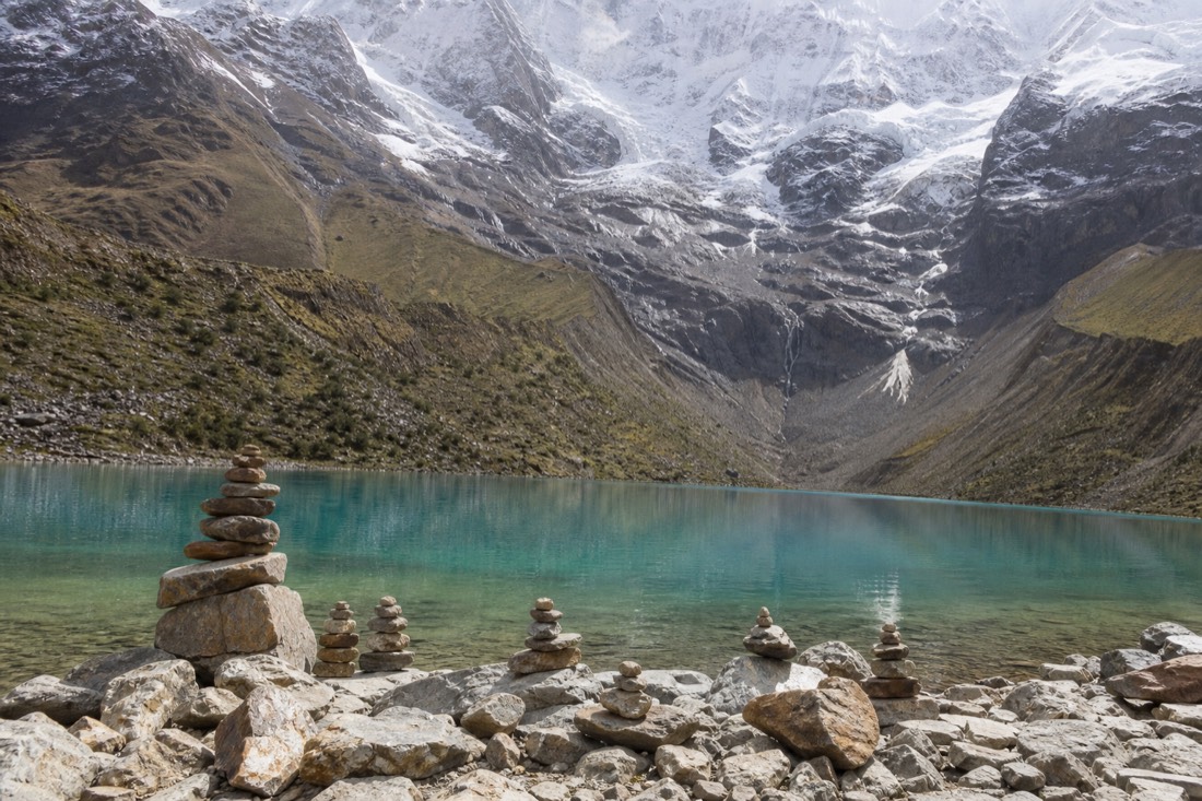 Glacier and grey moraine rising above the turquoise water of Humantay Lake with stacked stone cairns on the shore, no people