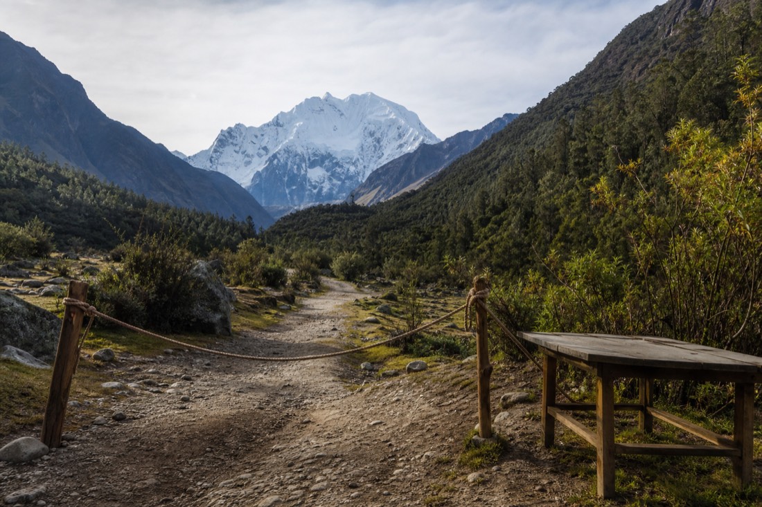 A simple rope barrier across a dirt road beside a wooden folding table at an Andean trail checkpoint below Soraypampa, pine slopes and a snow peak behind in early light, no people