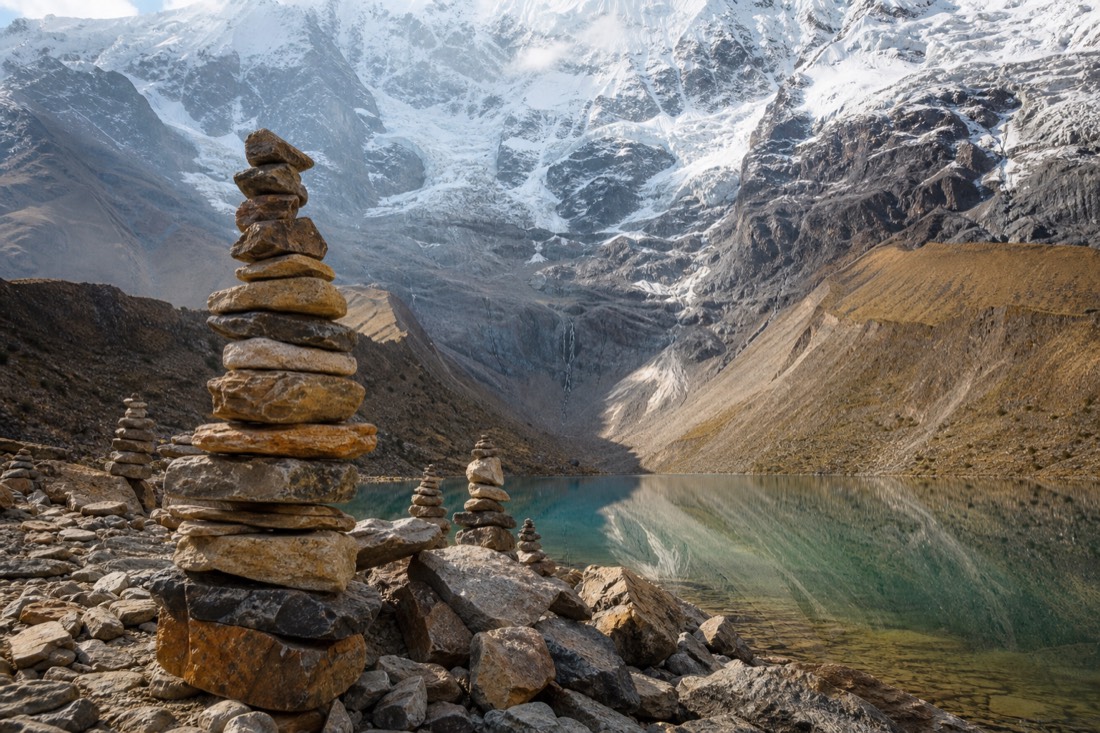 Cluster of stacked stone apachetas along the rocky shore of Humantay Lake with the glacier reflected in still turquoise water, no people