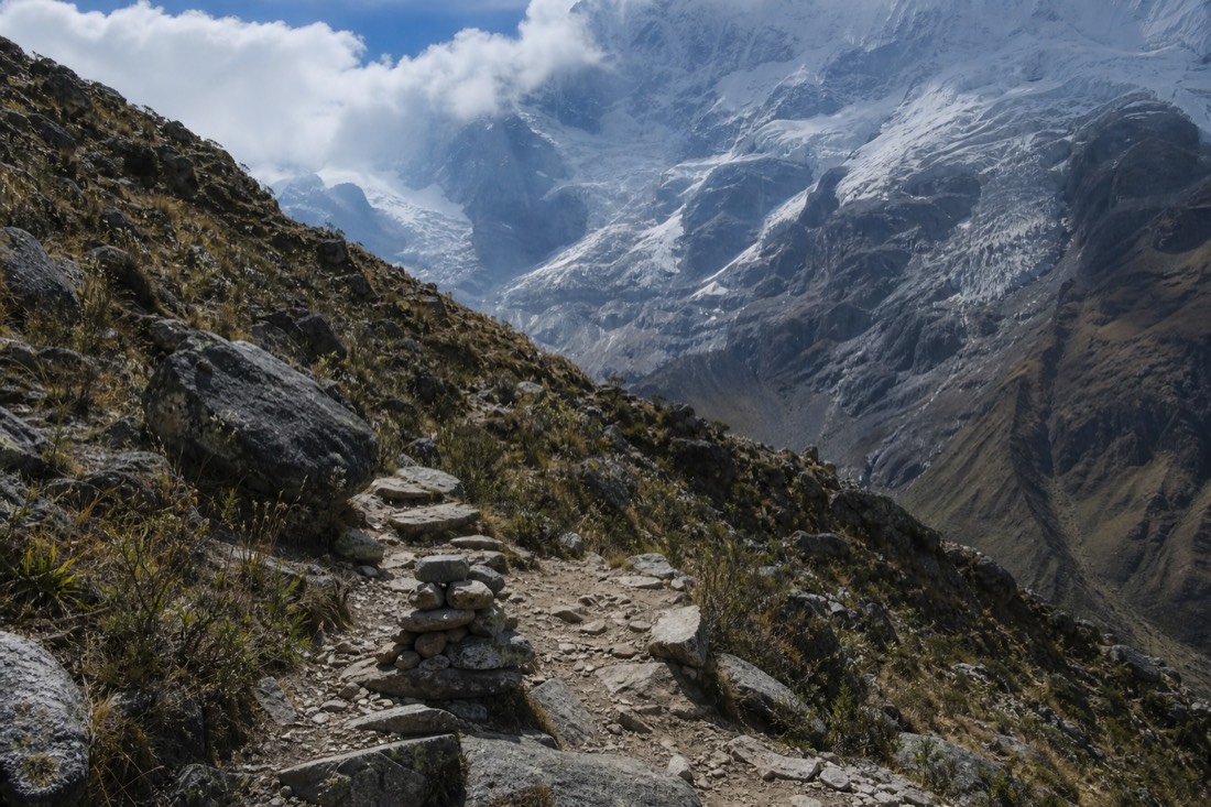 Rocky switchback trail climbing above Soraypampa toward a hanging glacier with scattered stone cairns and no people in frame