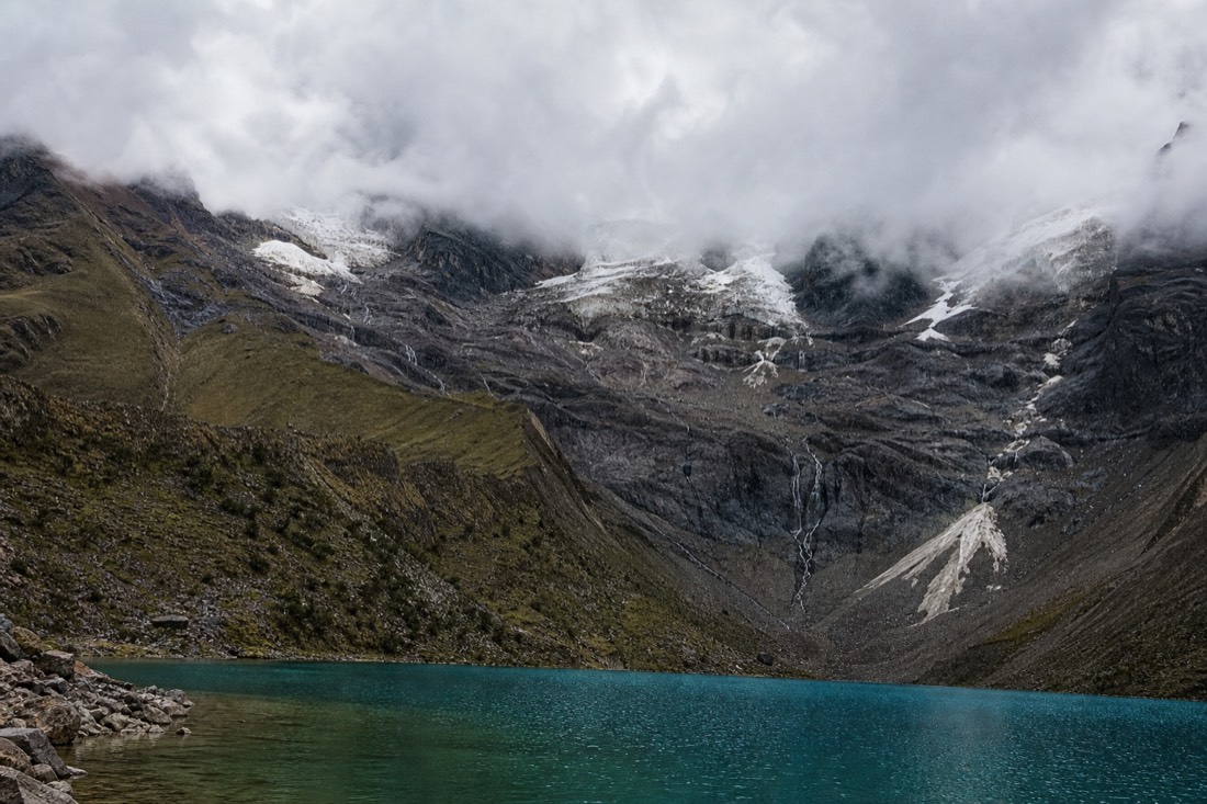 Humantay Lake under heavy grey cloud with muted slate-blue water and the glacier partly hidden, moody high-altitude scene with no people