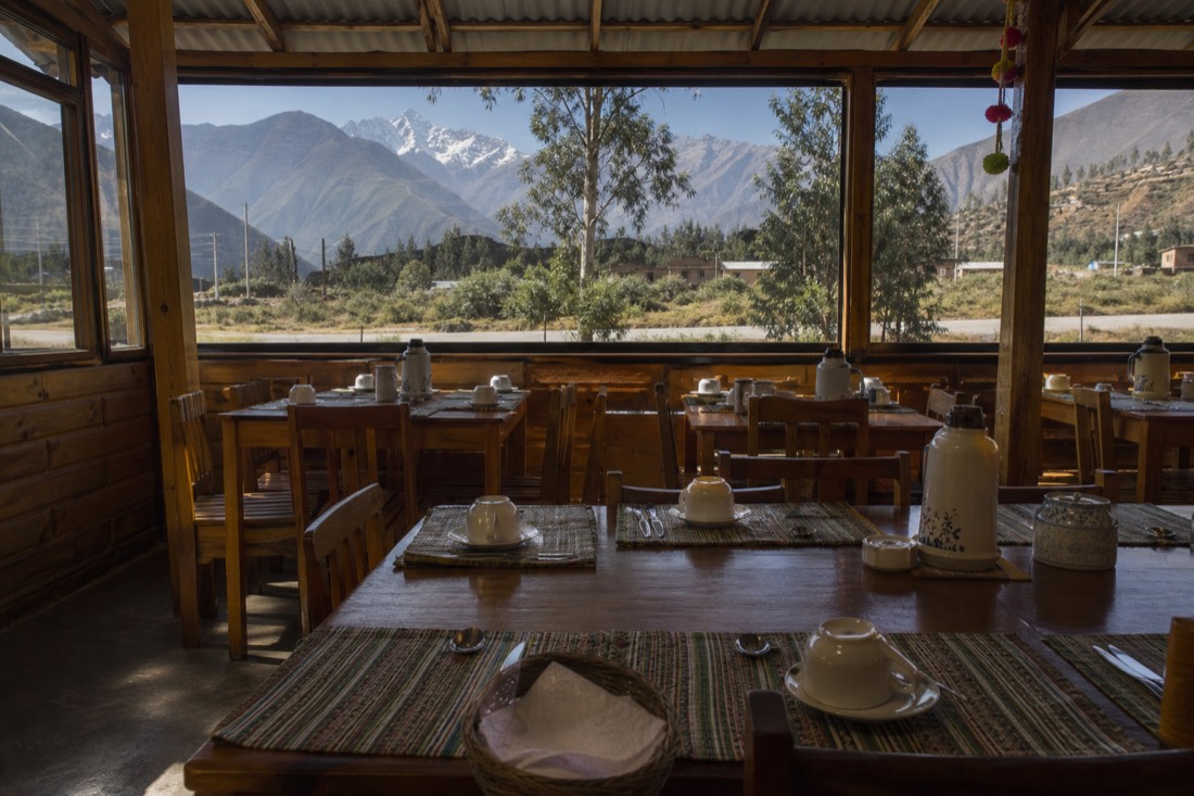 A simple roadside breakfast hall in the Andean village of Mollepata at first light, empty wooden tables set for a tour group, mountains framed in the window, no people