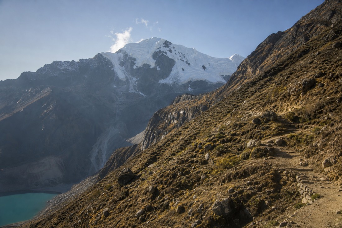 Steep rocky switchbacks climbing a treeless Andean slope toward Humantay in early morning light, a thin dusty trail, glacier peak above, no recognizable faces