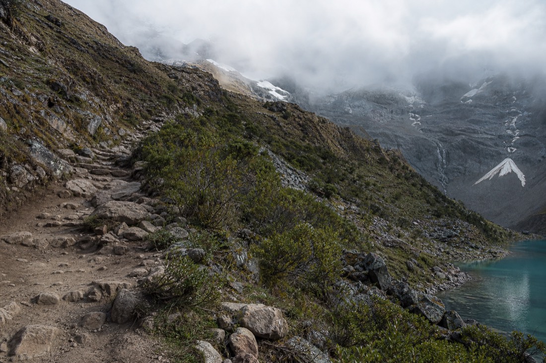 Line of the rocky switchback trail climbing toward Humantay Lake with cloud spilling over the ridge above, no people