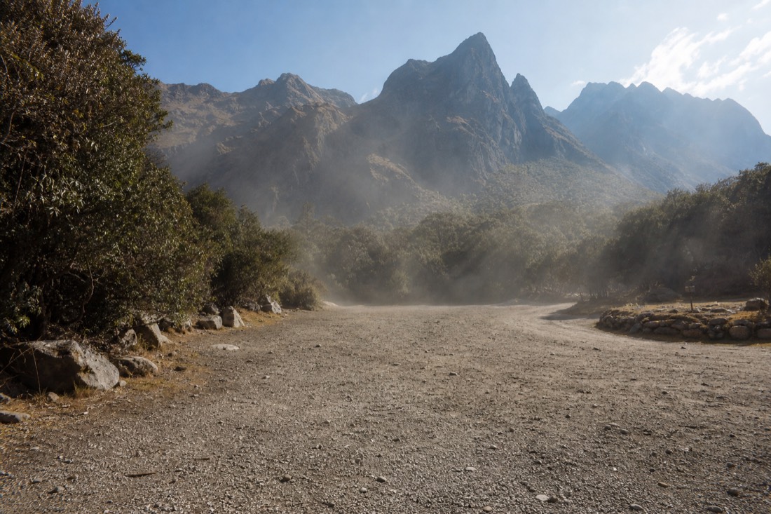 Empty gravel parking area at Soraypampa in mid-afternoon light with lingering dust and dark mountains behind, no vehicles or people
